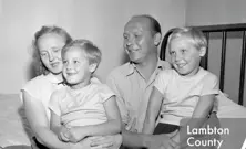 A black-and-white photograph of a smiling family of four, including two young children, sits closely together on a bed in a modest room.