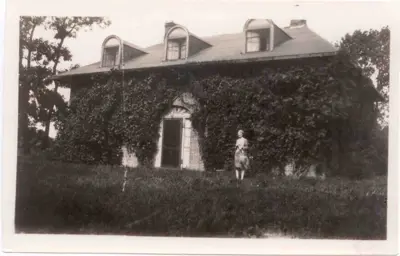 A black-and-white photograph shows a brick house covered in ivy with three dormer windows, and a woman standing in the grassy yard in front of the entrance.
