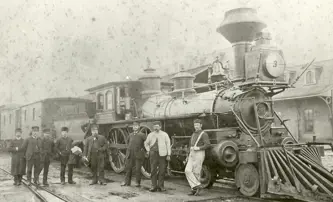 A vintage photograph shows a group of men standing beside a steam locomotive numbered 281 at a train station, with train cars extending into the distance.
